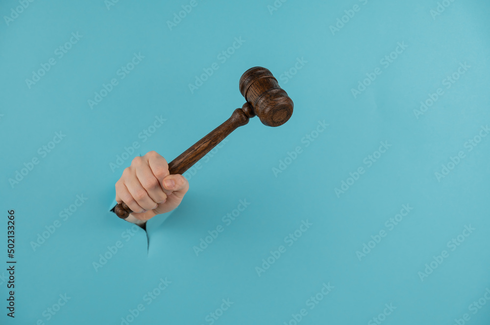 A woman's hand with a wooden judge's gavel sticks out of a hole in a blue background. Copy space. 