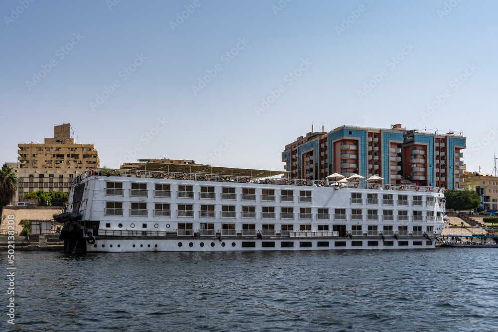 The tourist ship is moored at the river bank. Cabin windows and railings are visible. On the upper deck there is an awning, umbrellas. Ripples on the water. City houses against the blue sky. Egypt