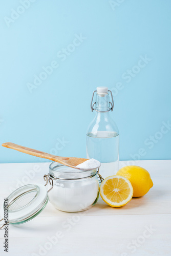 Open jar of baking soda with a wooden spoon on top, vinegar, cut lemon, on a blue background. The concept of organic removing stains on clothes