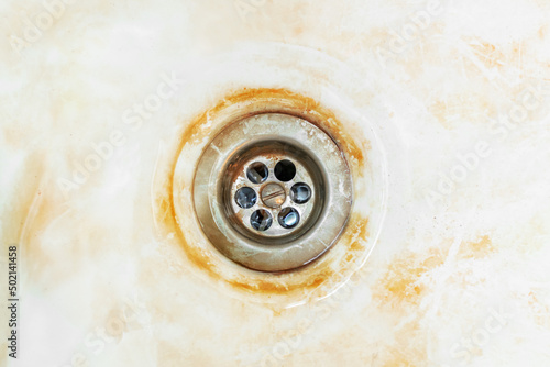 An old rusty bathtub with a metal drain hole. Dirty cracked unclean surface of the bath or sink with red rust stain, close-up. Corrosion, unsanitary concept.