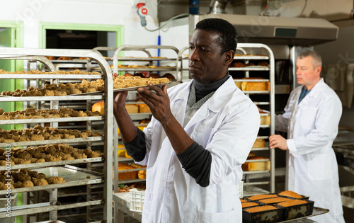 Skilled African American baker arranging trays with freshly baked bakery products on trolley..