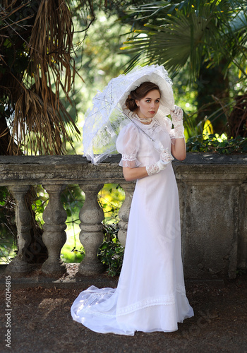 Obraz na plátně Young woman in white edwardian style dress and white big hat standing in old abandoned castle