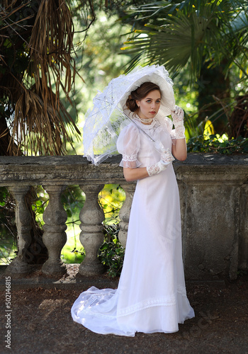 Young woman in white edwardian style dress and white big hat standing in old abandoned castle. Portrait of aristocratic girl. photo