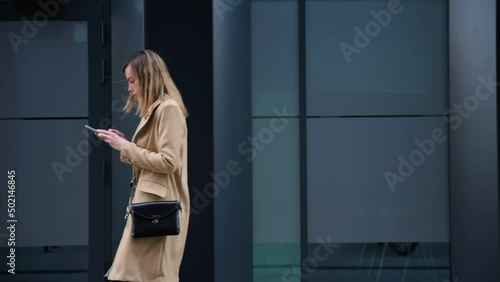 Stylish millenial woman in trench walks alone at city street and use smartphone photo