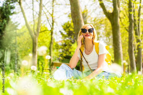 Tourist blonde woman calling her friends with phone, wearing a hat and sunglasses sitting on the grass in spring next to daisies in a park in the city