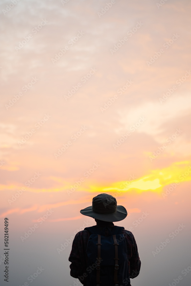 A man wearing a backpack in a trekking hat is standing in the evening watching the sun set in the clouds.silhouette travel and copy space.
