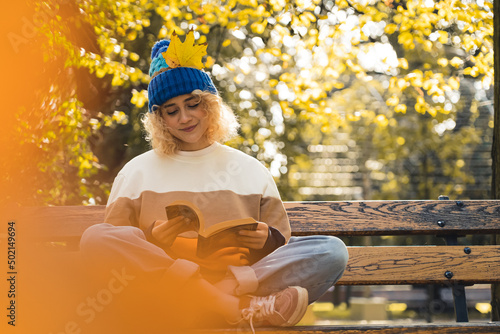 Beautiful young caucasian blonde girl in a knitted hat with a yellow leaf reading a book or the Bible on a bench in the autumn park. . High quality photo