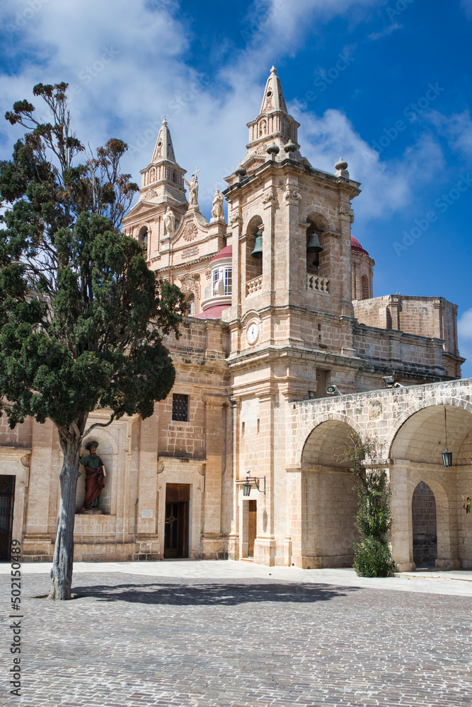Old church in Malta with big towers on a perfect blue sky background with a big tree in front casting its shadow over the yard creating a peaceful vacation scene