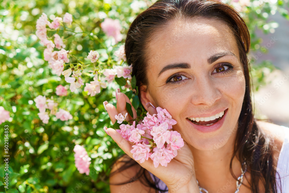 Close-up portrait beautiful woman in park with blooming small pink roses. Romantic young brunette in white T-shirt poses against the backdrop of spring flowering bushes. Smiling happy caucasian girl. Close-up portrait beautiful woman in park with blooming small pink roses. Romantic young brunette in white T-shirt poses against the backdrop of spring flowering bushes. Smiling happy caucasian girl.