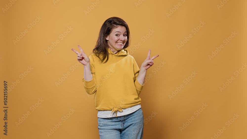 Portrait of casual smiling woman showing peace sign hand gesture in studio. Smiling girl showing victory sign celebrating on yellow background. Happy young adult making v sign with fingers. Portrait of casual smiling woman showing peace sign hand gesture in studio. Smiling girl showing victory sign celebrating on yellow background. Happy young adult making v sign with fingers.