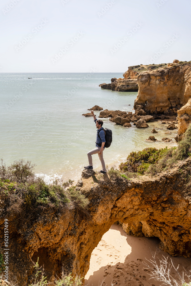 Carefree man with hand raised standing on rock bridge in front of sea Carefree man with hand raised standing on rock bridge in front of sea