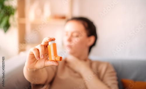 A girl suffering from an asthma attack due to allergies shows an inhaler, and coughs in the background, sitting on the couch. the concept of combating allergies. selective focus