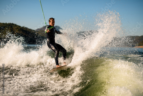 active man holds rope with handle and riding on wakesurf board on splashing river wave.