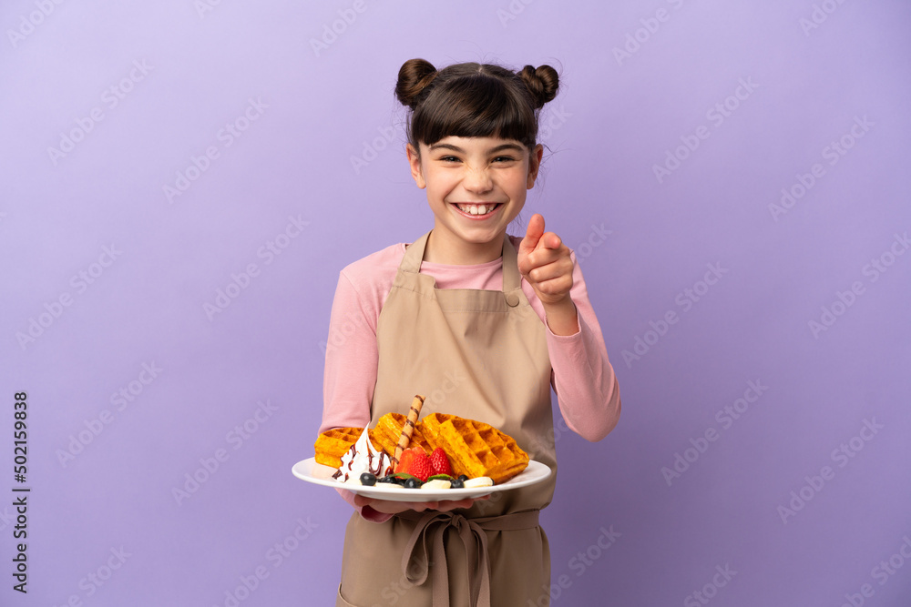 Little caucasian girl holding waffles isolated on purple background surprised and pointing front