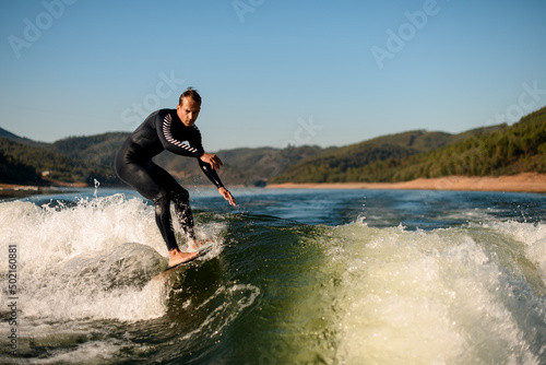 flexible man in wetsuit on wakesurf skilfully riding on splashing river wave