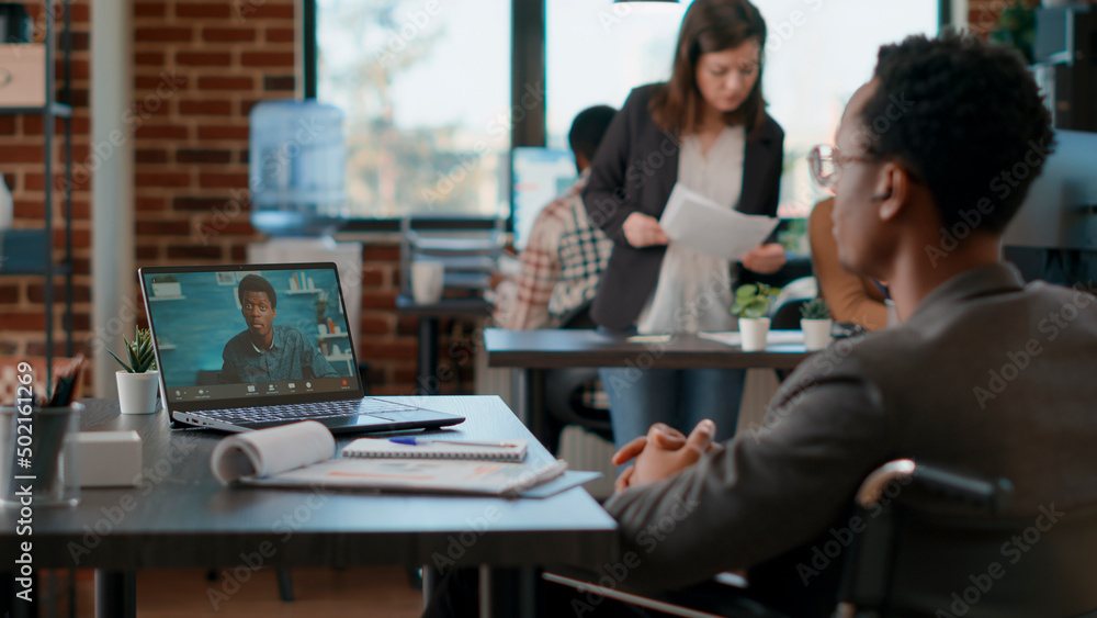 Fototapeta premium Office worker in wheelchair meeting with colleague on videocall conference, talking about business project. Professional employee using online teleconference call to have conversation on laptop.