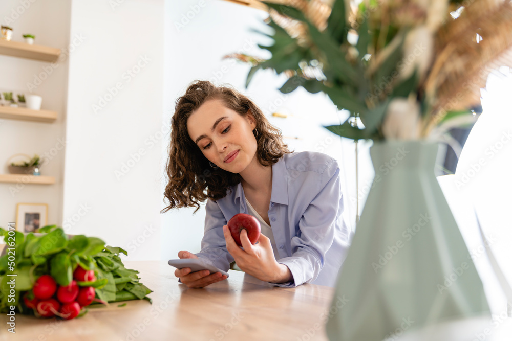 Young woman checking calories of apple on mobile phone in kitchen Young woman checking calories of apple on mobile phone in kitchen