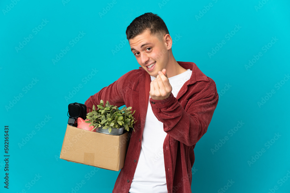 Young caucasian man making a move while picking up a box full of things isolated on blue background making money gesture Young caucasian man making a move while picking up a box full of things isolated on blue background making money gesture
