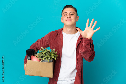 Young caucasian man making a move while picking up a box full of things isolated on blue background counting five with fingers