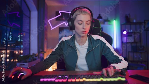 Gaming at Home: Female Gamer Putting on Headphones and Playing Online Video Game on Computer. Stylish Young Woman with Short Hair Enjoying Online Multiplayer Tournament. POV from Screen Perspective.