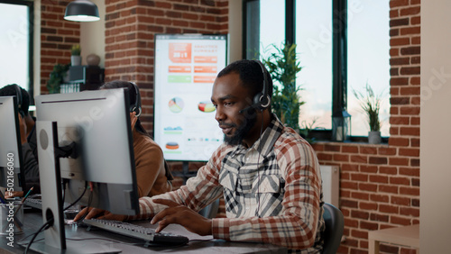 Young man using headphones at call center to help people with telemarketing problems. Helpdesk employee working at customer support service, giving assistance to business clients.