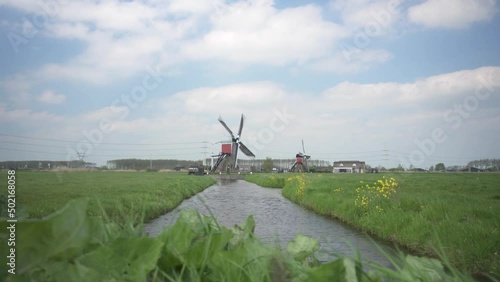 Windmills In The Farm With Green Grass And Stream In Foreground In Leiderdorp, Leiden, Netherlands. - wide photo