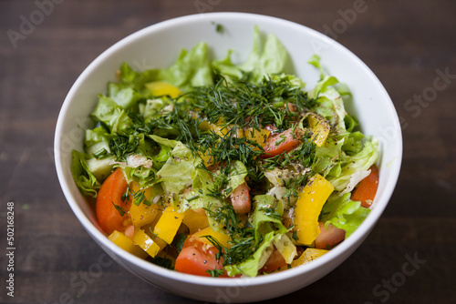 Closeup of chopped organic garden vegetables in a white bowl: tomatoes, cucumbers, yellow pepper, lettuce leaves and dill. Healthy nutrition. Cook at home.