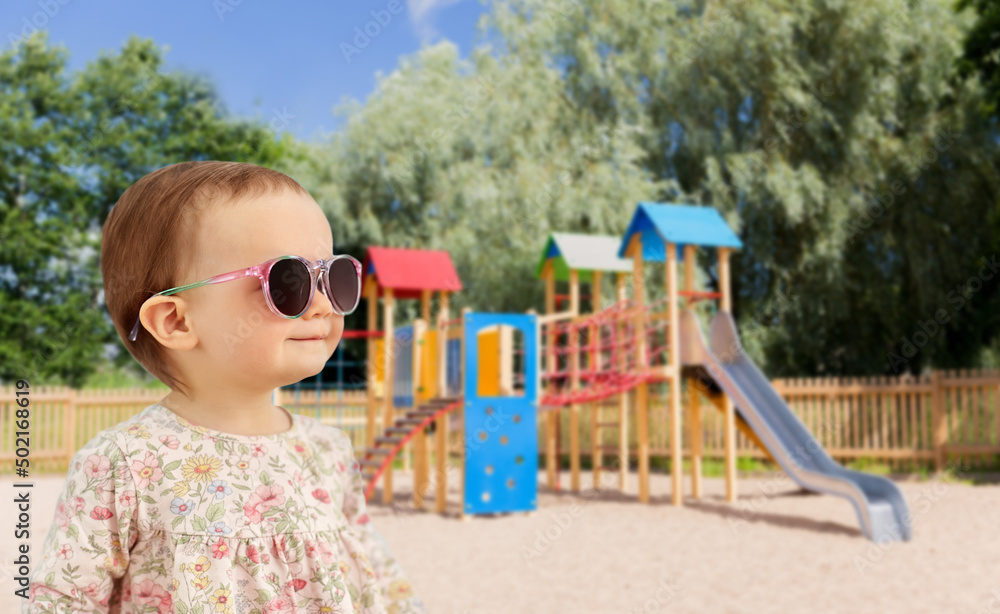 childhood, summer and people concept - happy little baby girl in sunglasses over children's playground background