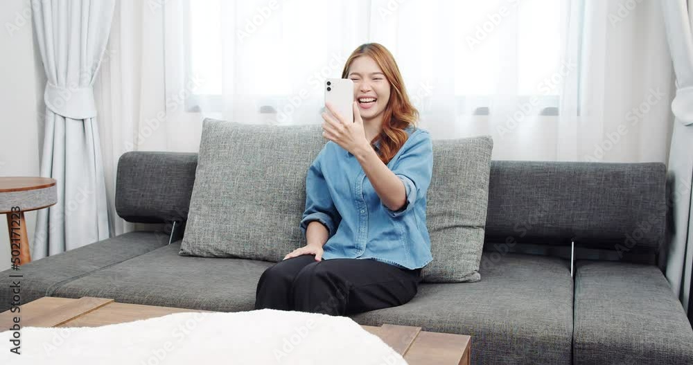 Charming Asian girl sitting on the couch uses video calls online technology with a friend in the cozy living room on a vacation day. Charming Asian girl sitting on the couch uses video calls online technology with a friend in the cozy living room on a vacation day.