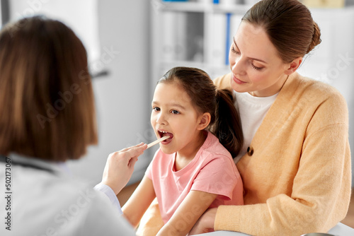 medicine  healthcare and pediatry concept - mother with little daughter and doctor with tongue depressor checking little girl patient s throat at clinic