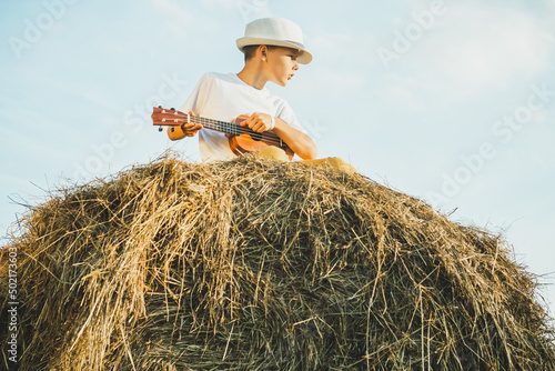 Portrait of barefoot boy in hat on haystack in field. Playing small guitar, ukulele. Light sunny day. Cheerful and music concept. Outdoor activity. Countryside