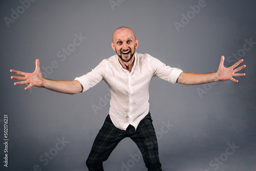 A bald guy in a white shirt and dark plaid pants throws up his hands on a gray background.