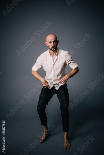 studio portrait of a bald guy with a beautiful beard barefoot in a white shirt and plaid pants dancing on a gray background