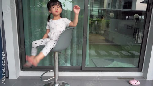 Asian child girl sitting on swivel chair ,Her face so bored and she want going outside with father,she piont hand to father outside the house want to go together. photo