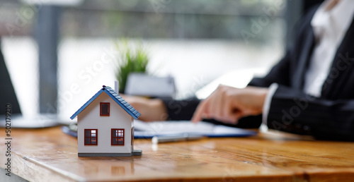 Businesswoman realtor taking notes and holding house model, sitting at desk with paper house model and keys, female real estate agent manager, preparing documents, mortgage and property.