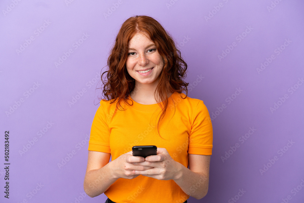 Teenager redhead girl over isolated purple background sending a message with the mobile Teenager redhead girl over isolated purple background sending a message with the mobile