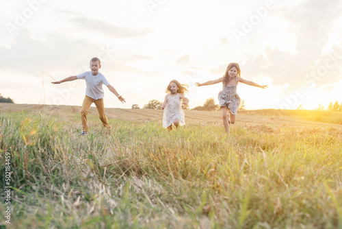 Portrait of three children playing game of catch, jumping and running on dry grass hay field paths in sunset. Trees and meadow on background. Looking around. Cloudy sunny sky. Haying time