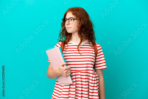 Student teenager redhead girl isolated on blue background looking to the side