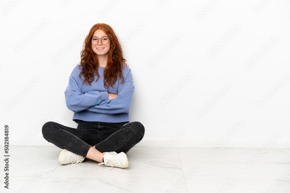 Teenager redhead girl sitting on the floor isolated on white background keeping the arms crossed in frontal position Teenager redhead girl sitting on the floor isolated on white background keeping the arms crossed in frontal position
