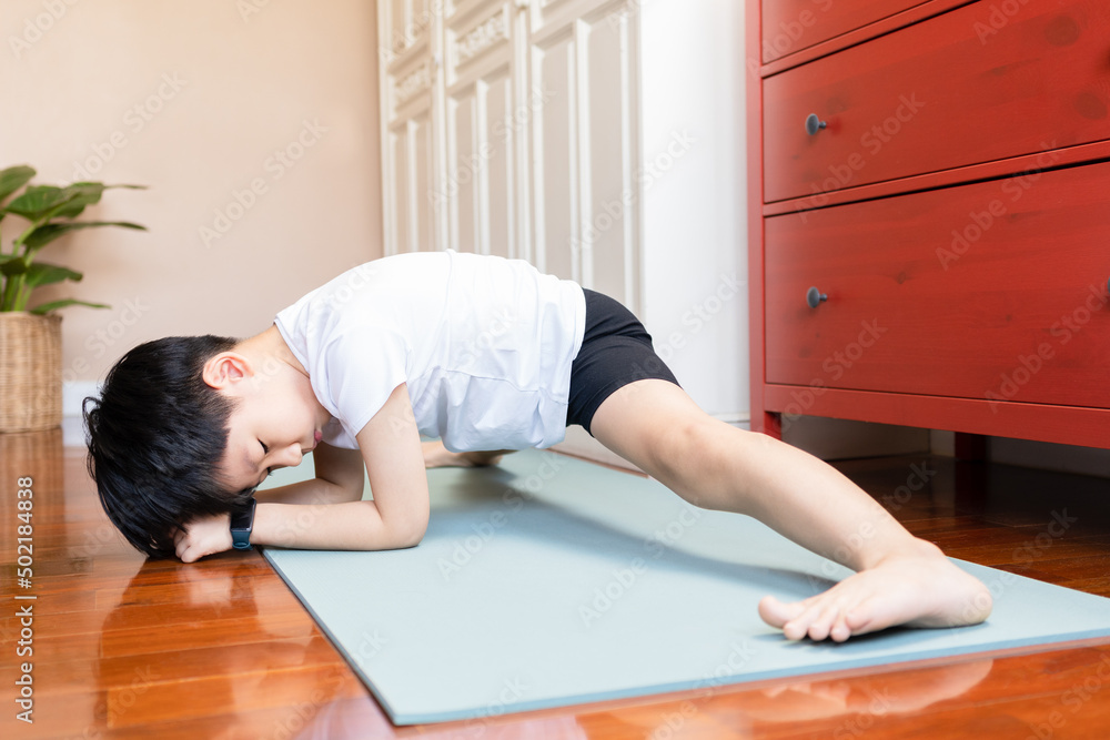 Fototapeta premium Cute Asian boy child practice leg stretch yoga pose and breathing on yoga mat in bedroom at home with concentration and discipline. Wellbeing, healthy and active lifestyle.
