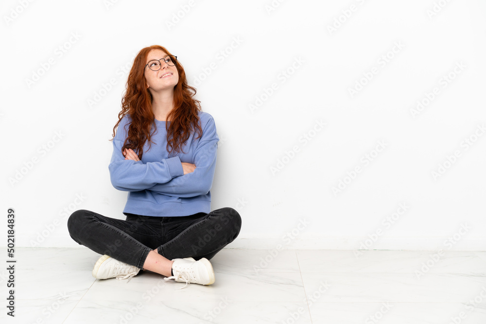 Teenager redhead girl sitting on the floor isolated on white background looking up while smiling Teenager redhead girl sitting on the floor isolated on white background looking up while smiling