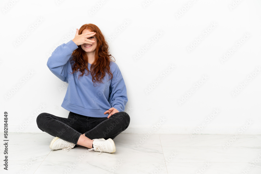 Teenager redhead girl sitting on the floor isolated on white background covering eyes by hands and smiling Teenager redhead girl sitting on the floor isolated on white background covering eyes by hands and smiling