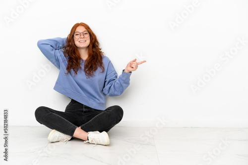 Teenager redhead girl sitting on the floor isolated on white background surprised and pointing finger to the side