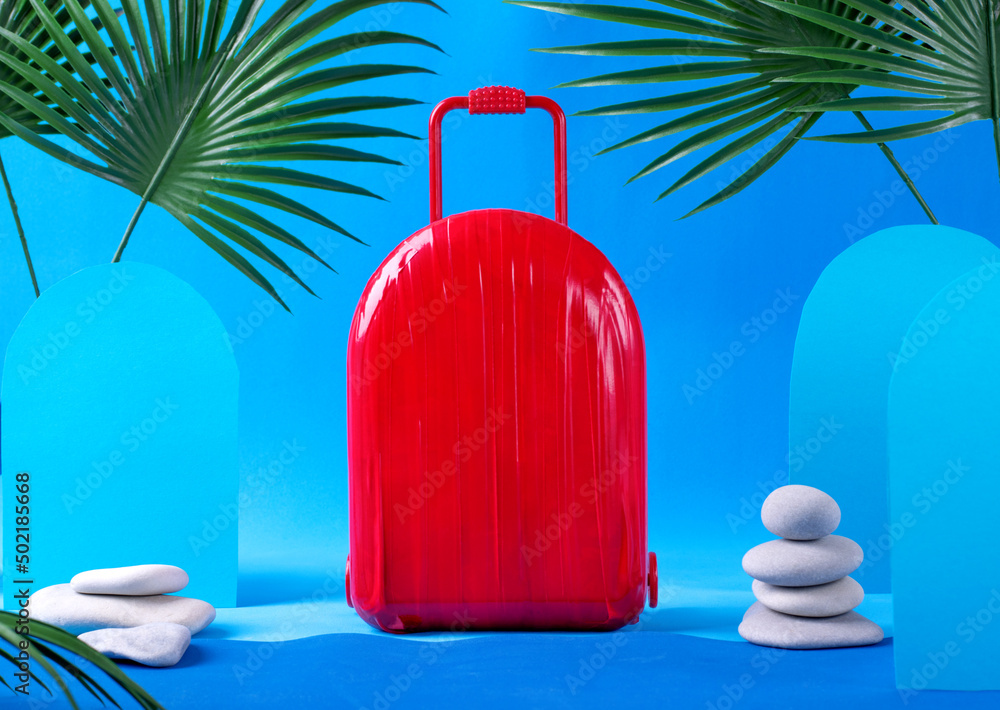 Red suitcase against the blue background with palm leaves and stones