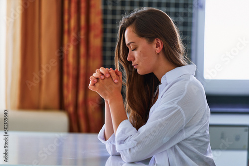Slika na platnu Lonely calm woman believer sits at table with praying hands alone at home and praying to God in silence for support and help