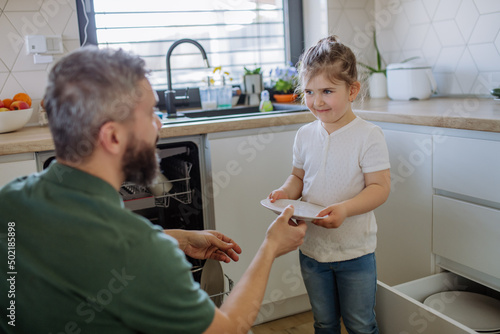 Little girl helping his father to unlad dishwasher in kitchen at home. photo