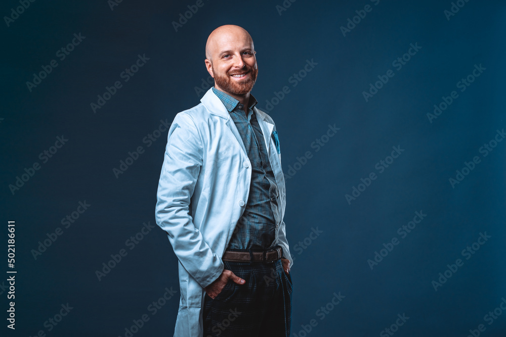 Photo of cheerful doctor with beard posing and smiling at camera with blue background and medical white coat Photo of cheerful doctor with beard posing and smiling at camera with blue background and medical white coat