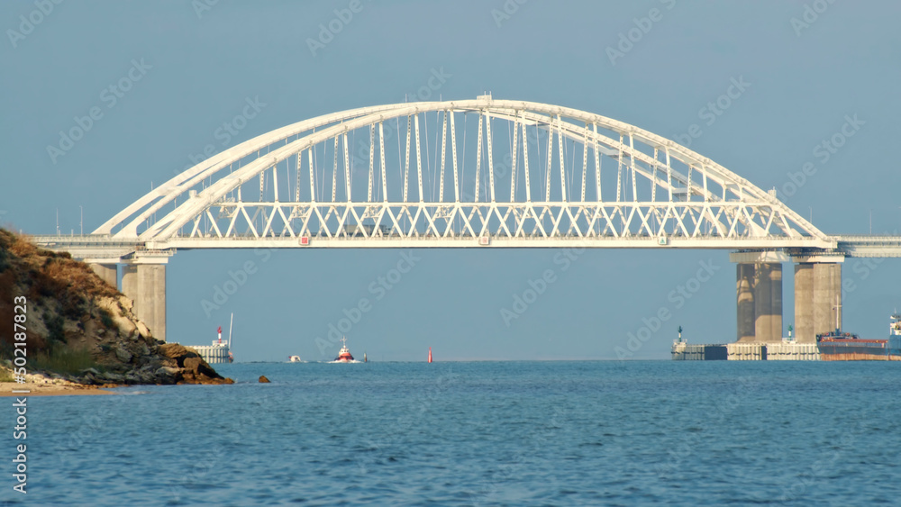 Fototapeta premium View of the beautiful large white bridge for cars moving above the calm sea and cargo ships against blue clear sky. Shot. Picturesque marine landscape
