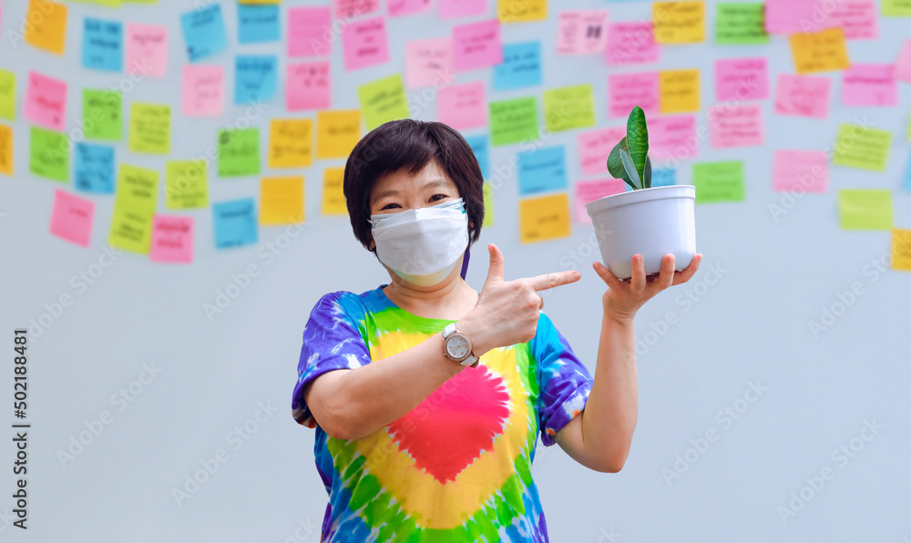 Asian cheerful woman in rainbow t-shirt with protective mask showing her little houseplant with blurred background of sticky notes on gray cement wall at home Asian cheerful woman in rainbow t-shirt with protective mask showing her little houseplant with blurred background of sticky notes on gray cement wall at home