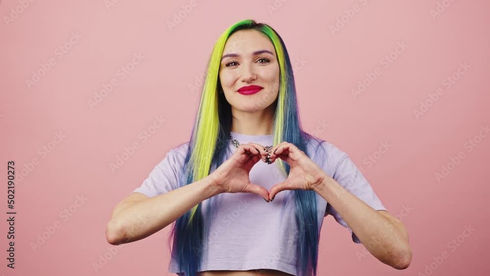 Young happy lady with bright colorful hairstyle showing heart gesture and sending love to camera, pink studio background Young happy lady with bright colorful hairstyle showing heart gesture and sending love to camera, pink studio background